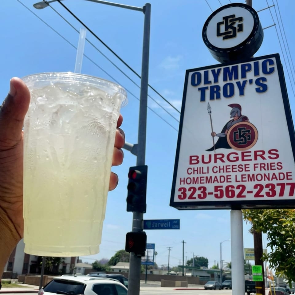 Hand holds an iced homemade lemonade beside an Olympic Troy’s sign advertising burgers, chili cheese fries, and lemonade.